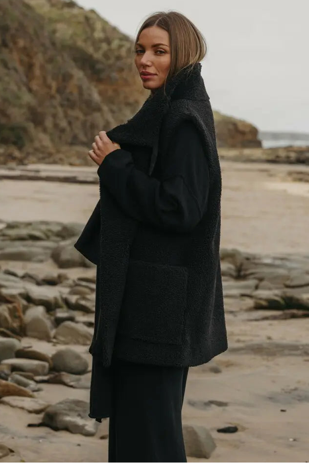 Woman in a black coat standing on a rocky beach with a scenic background