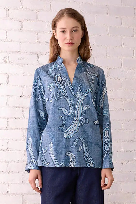 Woman wearing a blue patterned blouse and dark pants standing against a white brick wall.