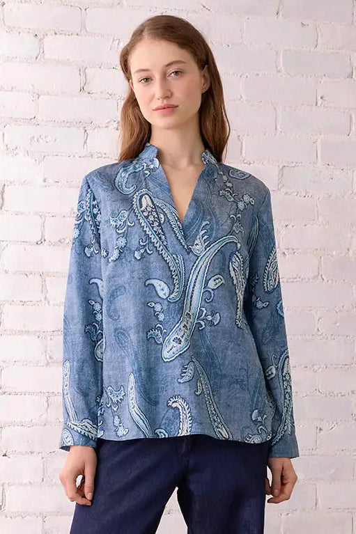 Woman wearing a blue patterned blouse and navy pants standing against a white brick wall.