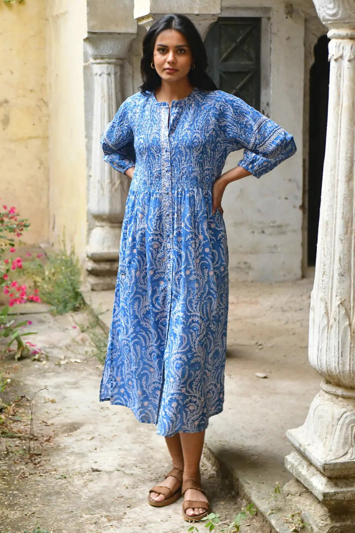 Woman in a blue dress standing in an outdoor setting with columns and plants.