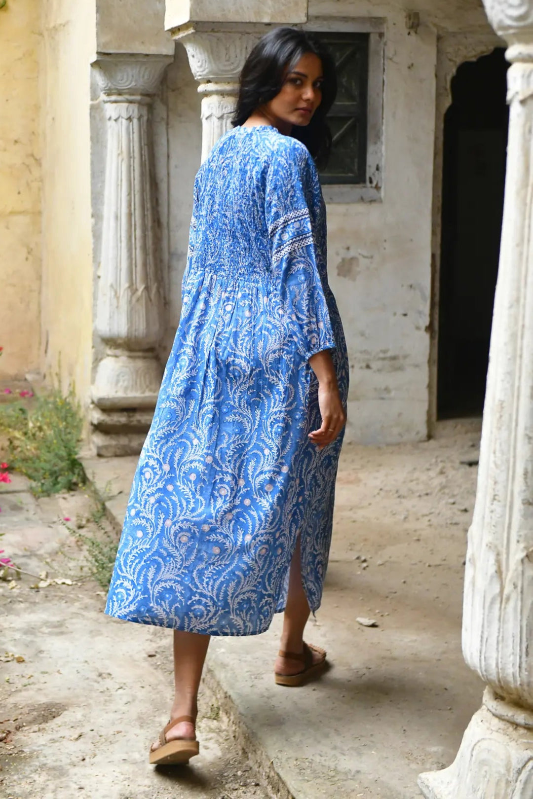 Woman in a blue patterned dress walking through an outdoor setting with columns and plants.