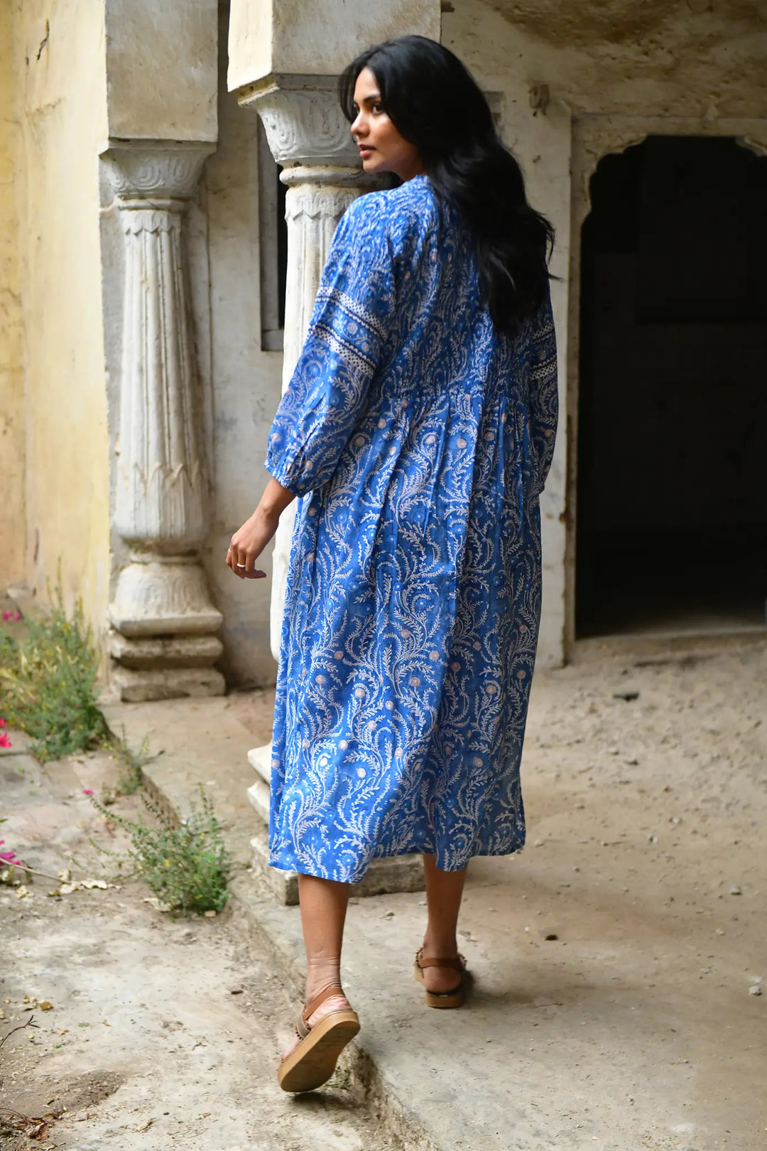 Woman in a blue patterned dress walking in an outdoor setting with stone architecture.