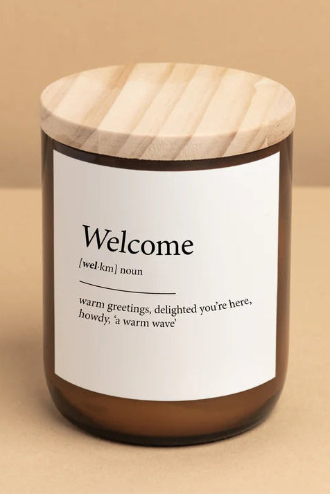 A soy candle in a clear glass jar with a wooden lid, labeled 'Welcome' with a definition below the word.