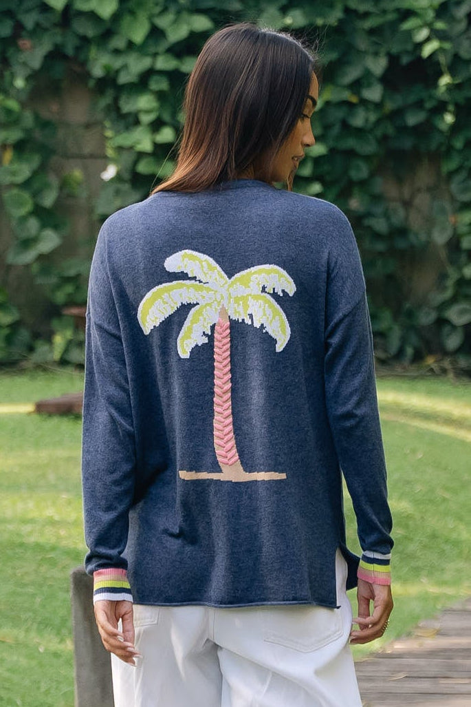 A woman viewed from behind walking on a wooden boardwalk in a garden, wearing a navy long-sleeve top with a large embroidered palm tree on the back and white wide-leg pants.