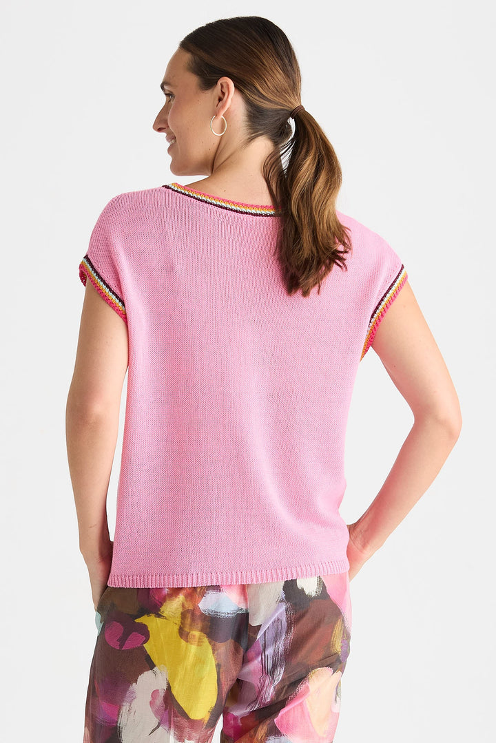 A woman shown from the back wearing a light pink short-sleeve knit top with thin multicolor striped trim along the neckline and sleeve cuffs, paired with floral print pants against a light gray studio background.