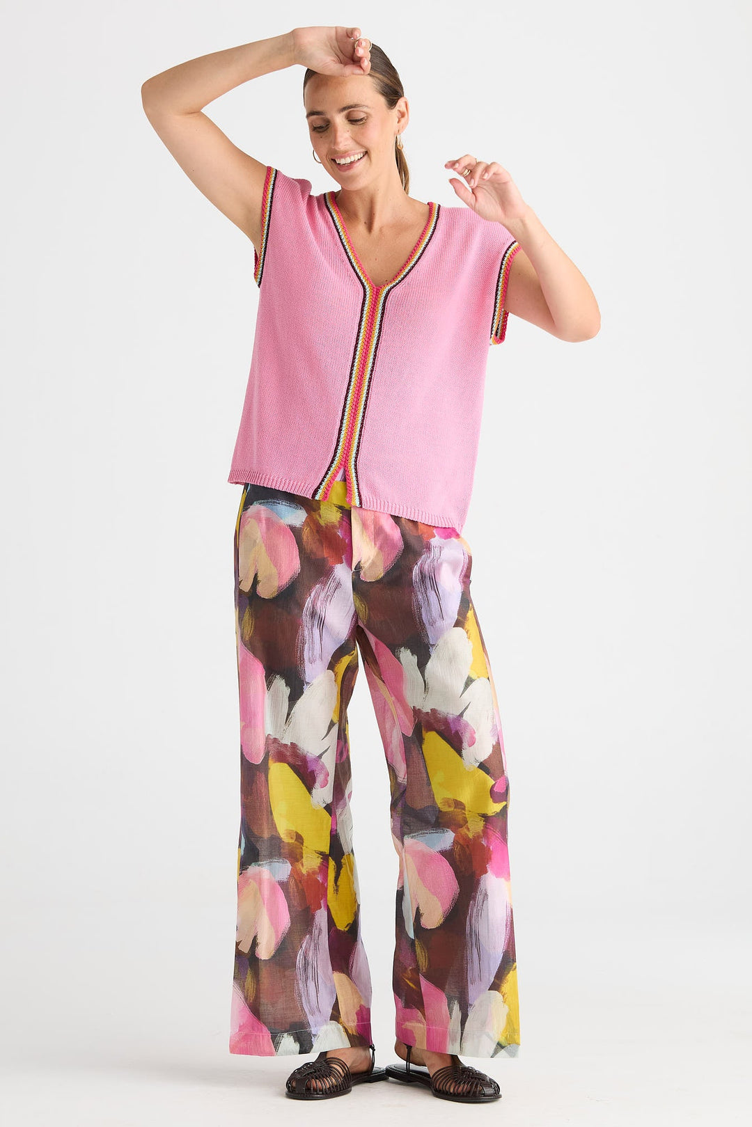 A woman modeling a sleeveless pink top with a colorful striped trim and matching wide-leg pants featuring a large abstract floral print in pink, yellow, brown, and white, set against a plain light gray background.
