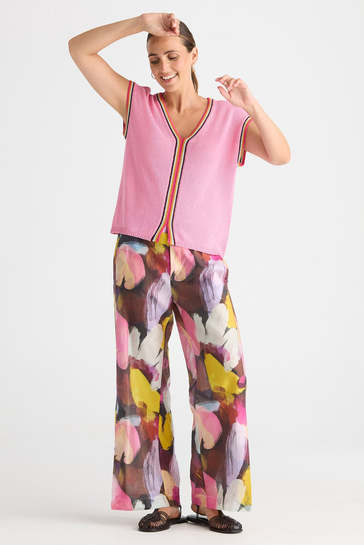 A woman modeling a sleeveless pink top with a colorful striped trim and matching wide-leg pants featuring a large abstract floral print in pink, yellow, brown, and white, set against a plain light gray background.
