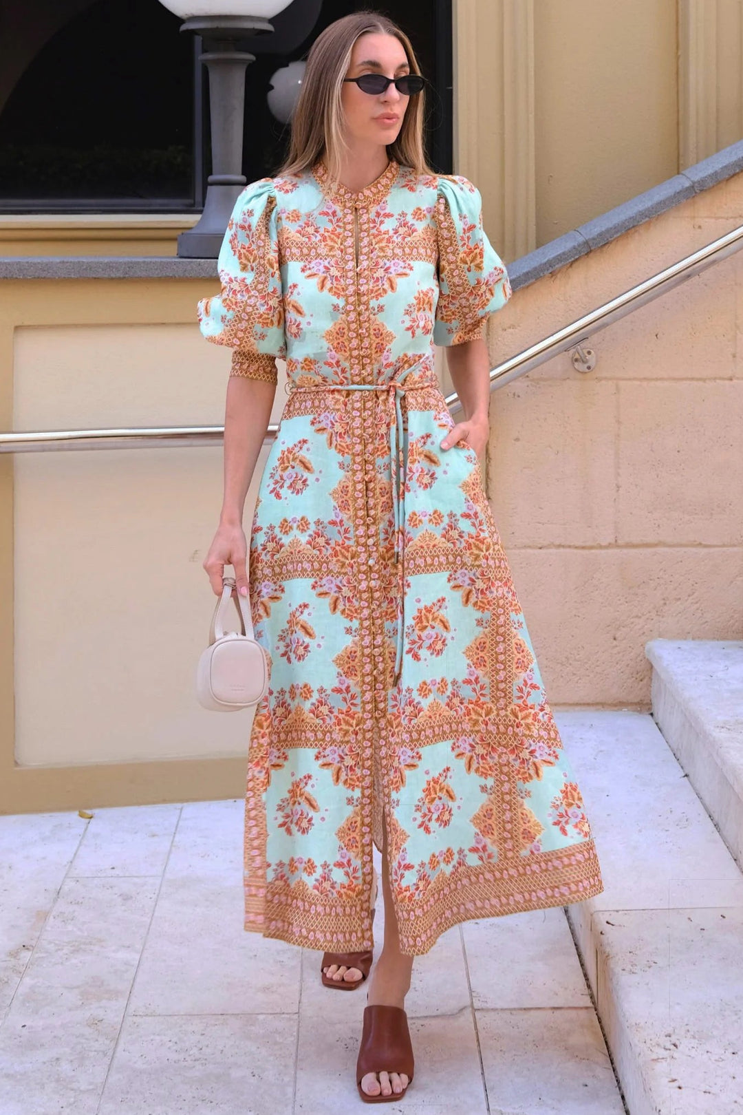 A woman wearing a mint green and orange patterned midi dress with puff sleeves and a tie waist, walking down outdoor stone steps while holding a small cream handbag.