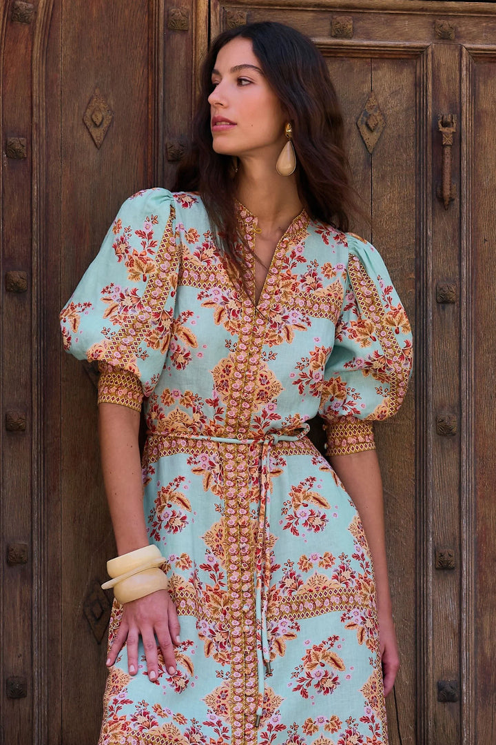 A woman wearing a light blue puff-sleeve dress with a brown and orange floral pattern, standing in front of a rustic dark wood door.