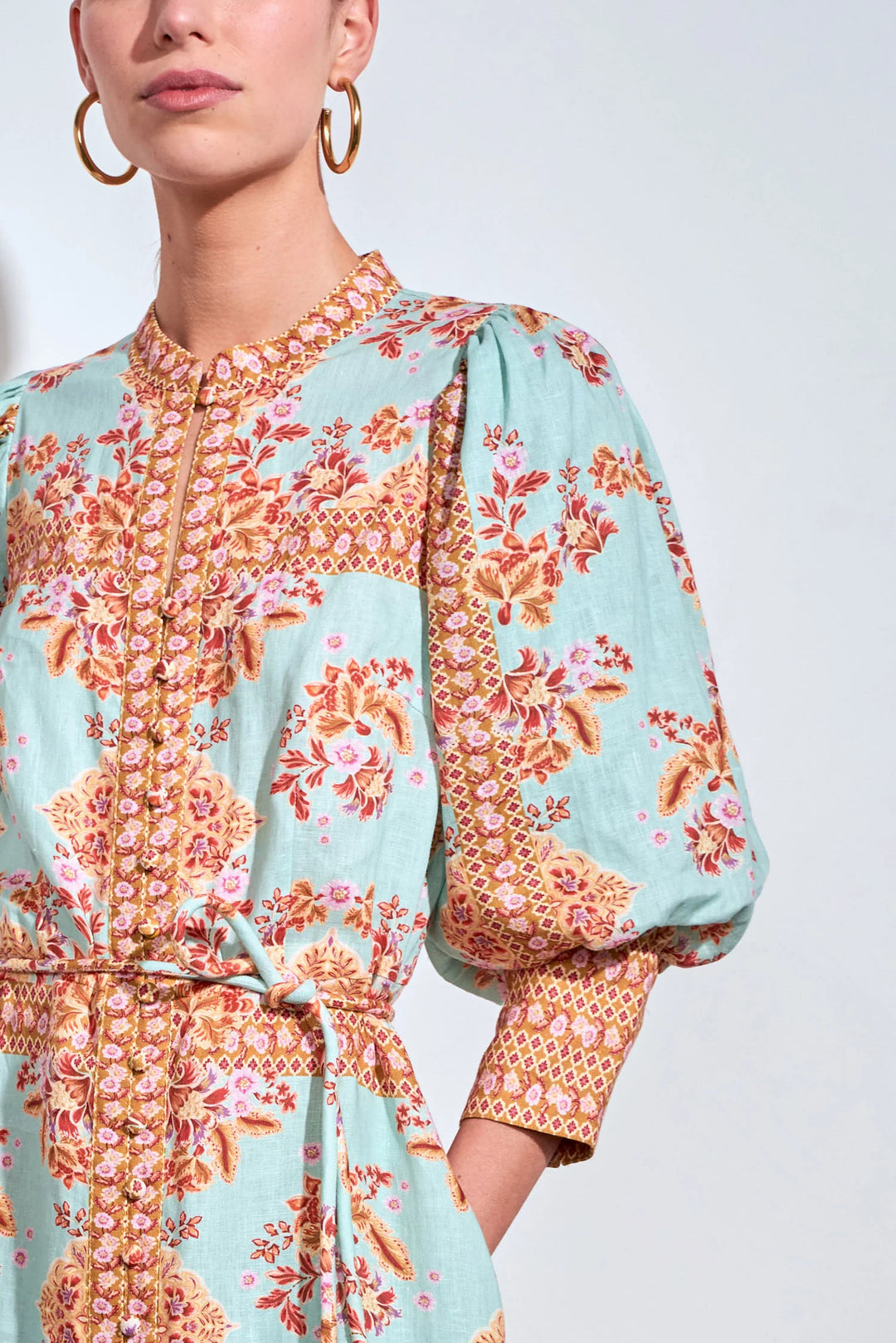 A close-up of a model wearing a light blue-green dress with a vibrant orange, pink, and gold floral pattern, featuring puff sleeves and a matching waist tie.