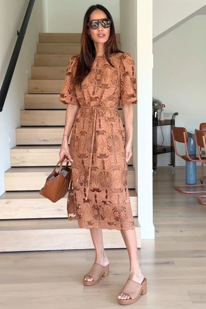 A woman models a rust-brown midi dress with a tonal floral lace pattern, puff sleeves, and a tie waist. She is standing in a modern home interior with light wood stairs and flooring, holding a matching brown handbag.
