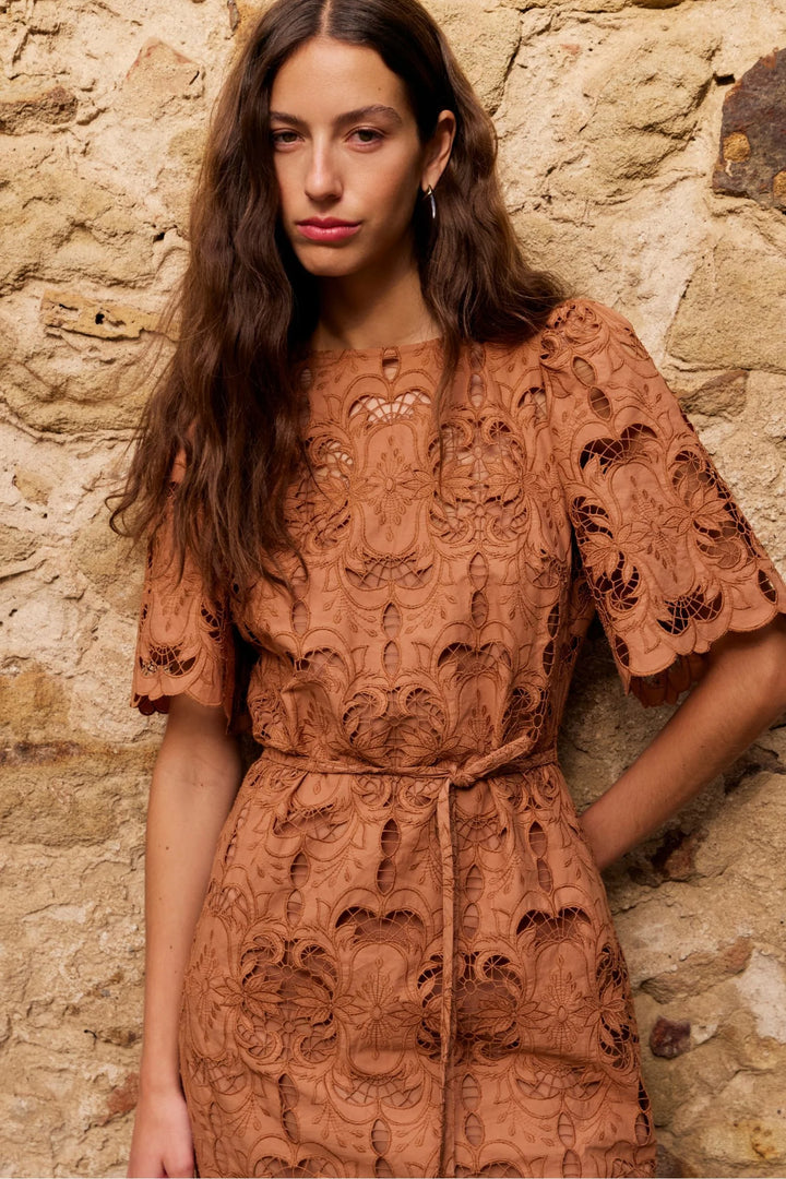 A woman wearing a rust-brown lace dress with a matching belt, standing against a rustic stone wall.