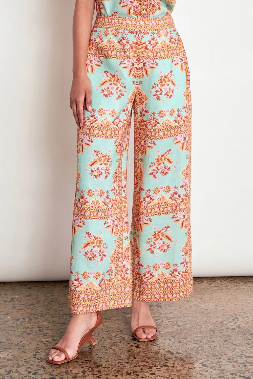 A model wears high-waisted, wide-leg cropped pants featuring a mint green base with orange and beige floral patterns, paired with brown heeled sandals against a neutral indoor background.