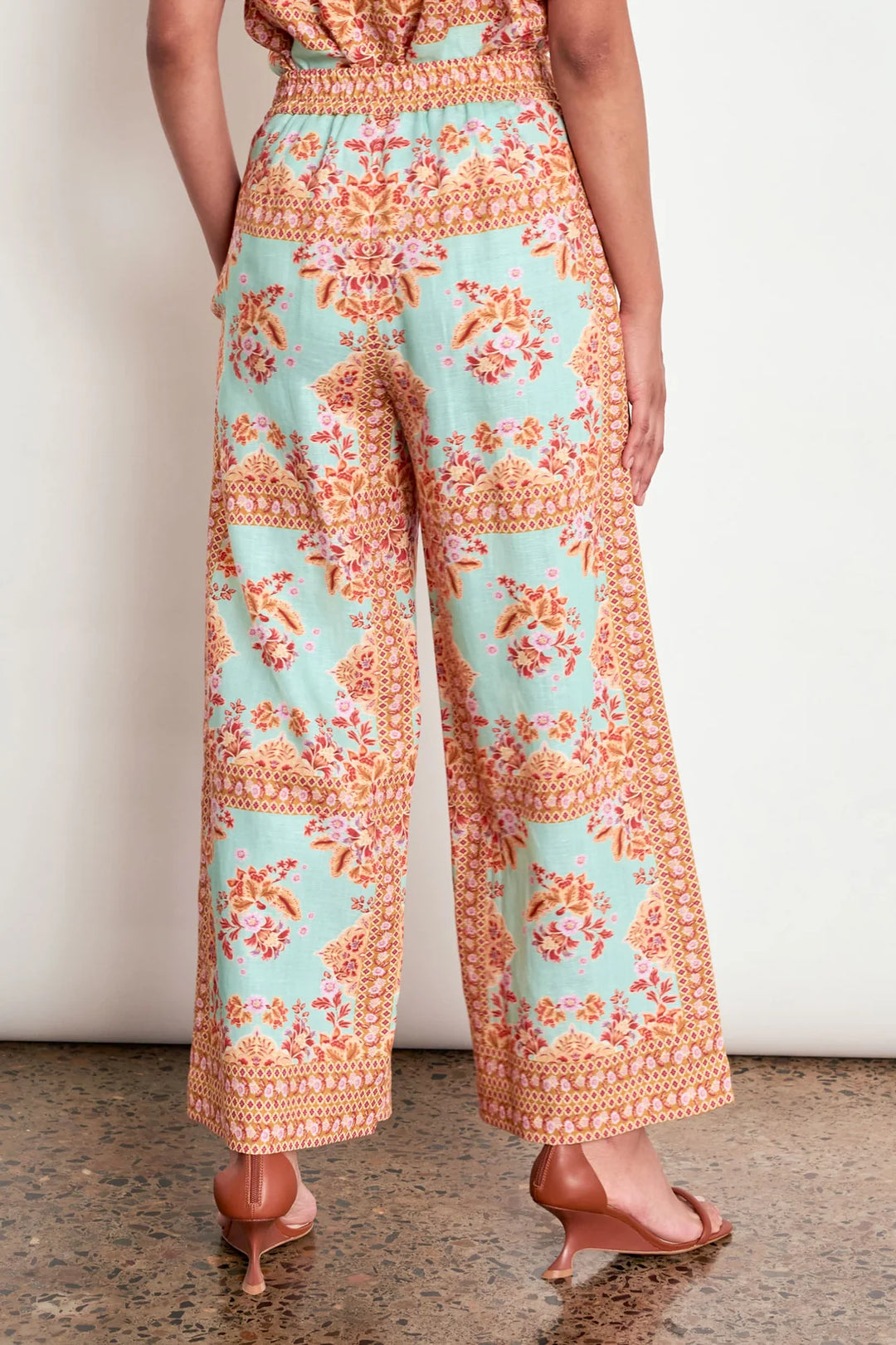 A person wearing wide-leg cropped pants with a mint green and orange floral patchwork print, paired with brown heeled sandals, shown from the back against a plain off-white wall.