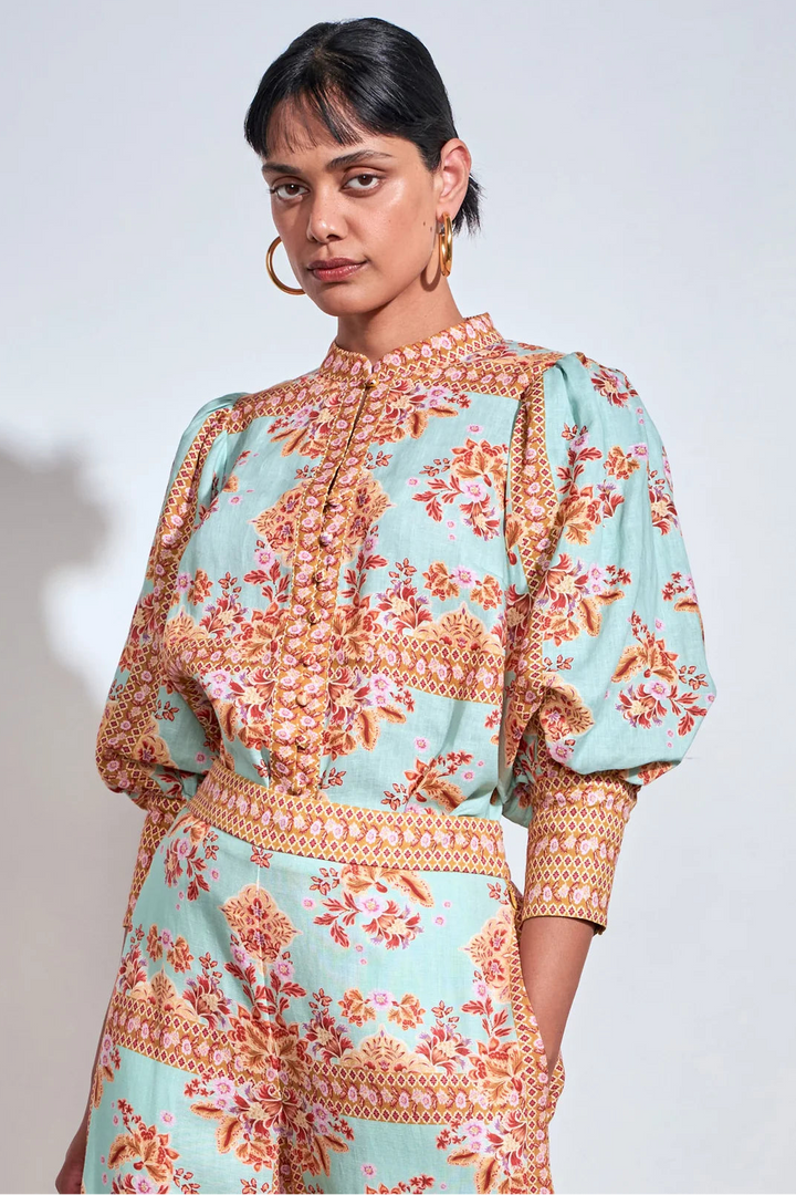 A woman modeling a light aqua blue jumpsuit with a vibrant orange, pink, and yellow floral and geometric pattern, featuring puff sleeves and a high collar.