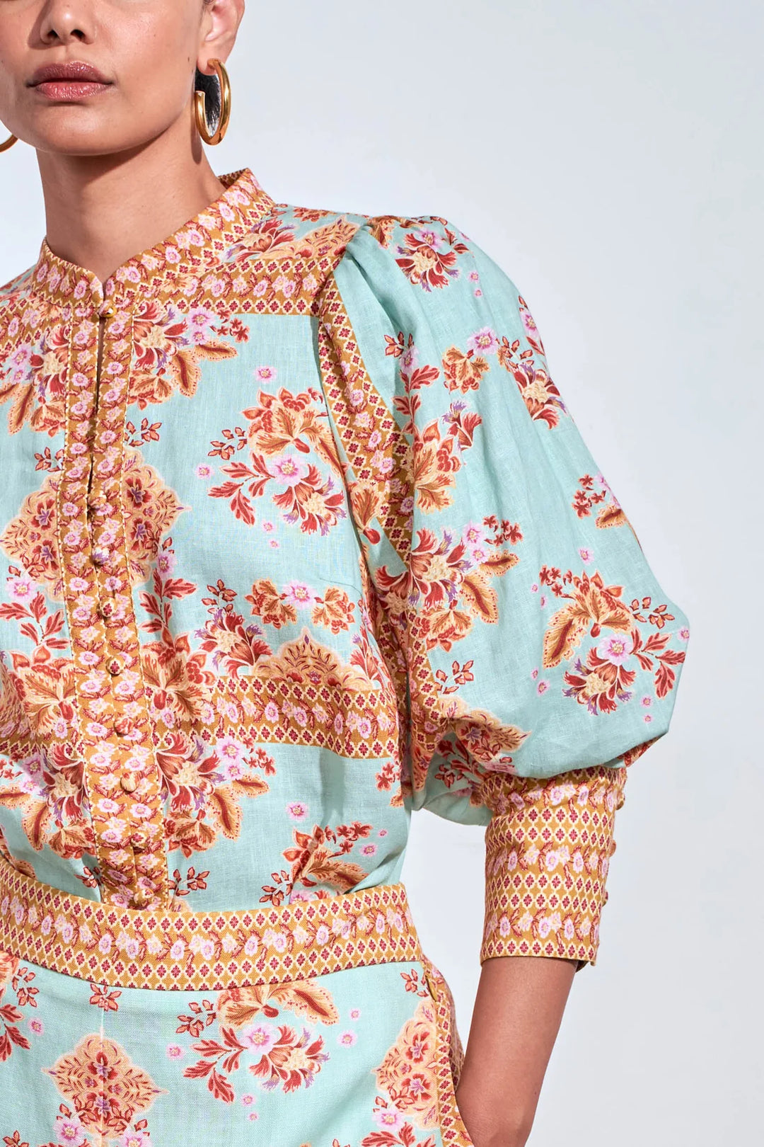 A close-up of a model wearing a light mint green printed top with orange and pink floral motifs and gold-toned embroidery. The top features a high collar, buttoned front, and voluminous elbow-length sleeves with patterned cuffs.