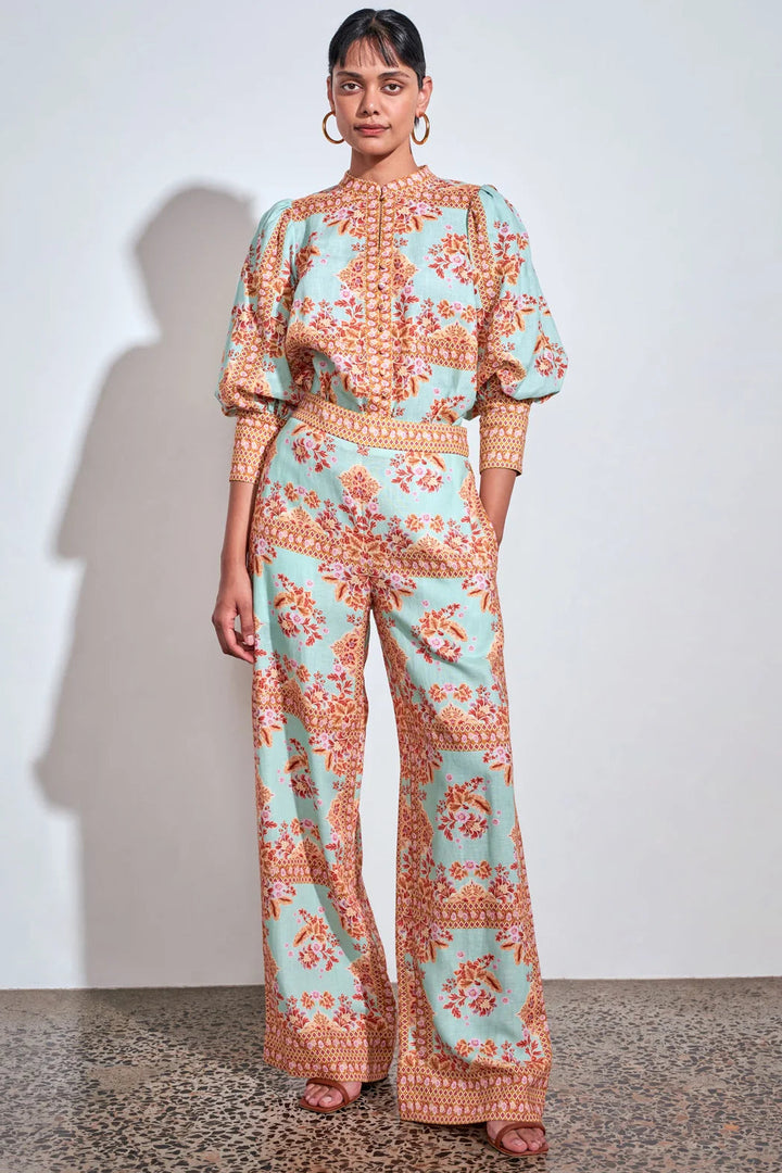 A woman modeling a light aqua and orange patterned jumpsuit with puff sleeves and wide-leg pants, standing against a plain white wall.