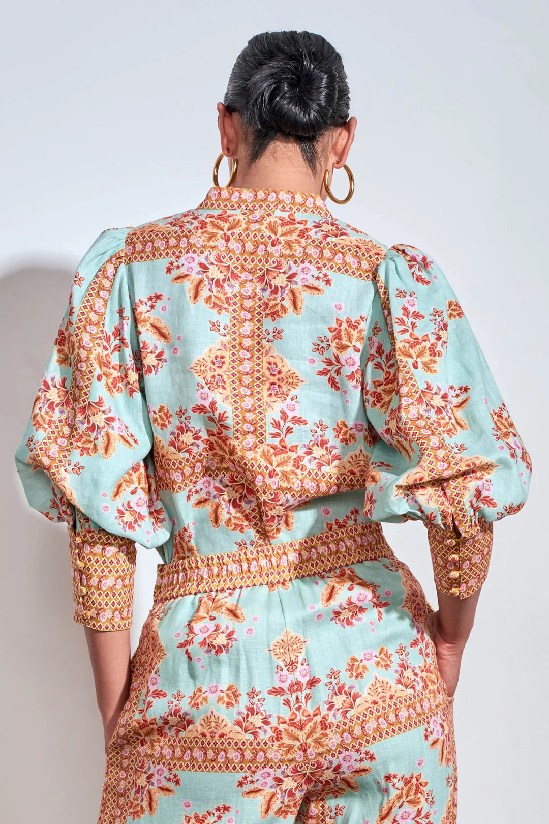 A woman viewed from the back wearing a mint green jumpsuit with a detailed orange and pink floral print and gold trim, paired with large gold hoop earrings.