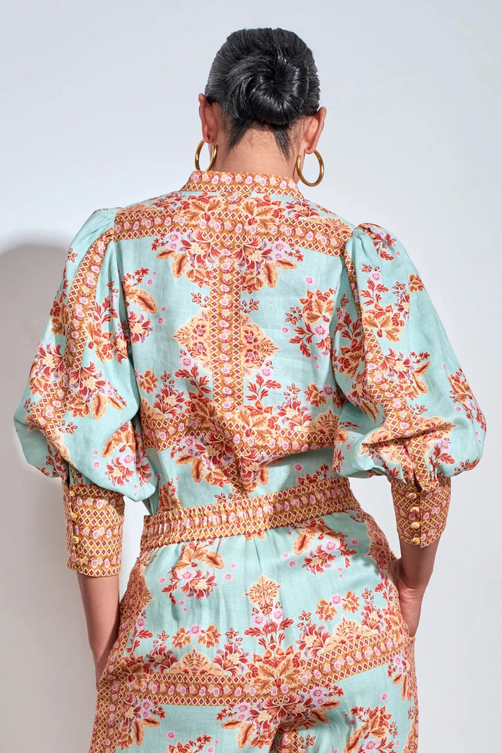 A woman viewed from the back wearing a mint green jumpsuit with a detailed orange and pink floral print and gold trim, paired with large gold hoop earrings.