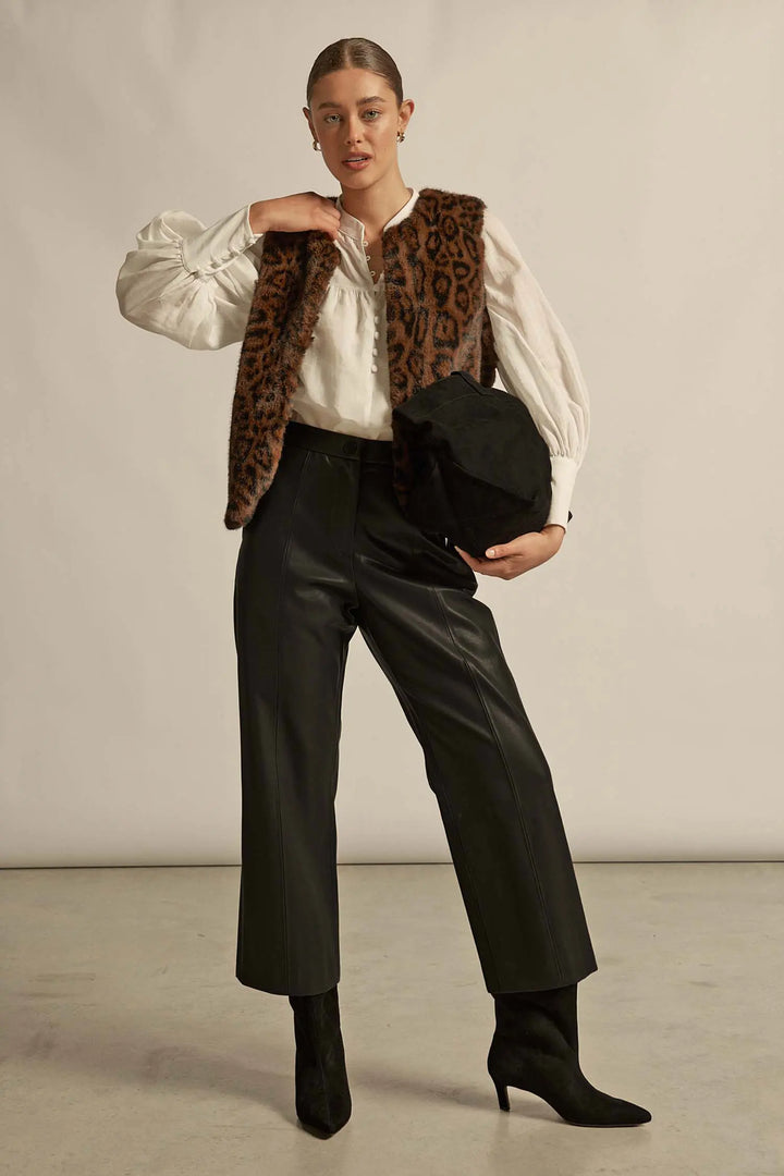 Person wearing a leopard print vest over a white blouse with black pants, holding a black hat.