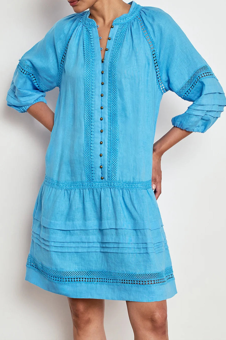 A woman wearing a light blue cotton tunic dress with embroidered details, a buttoned neckline, and a tiered skirt, shown against a plain light background.