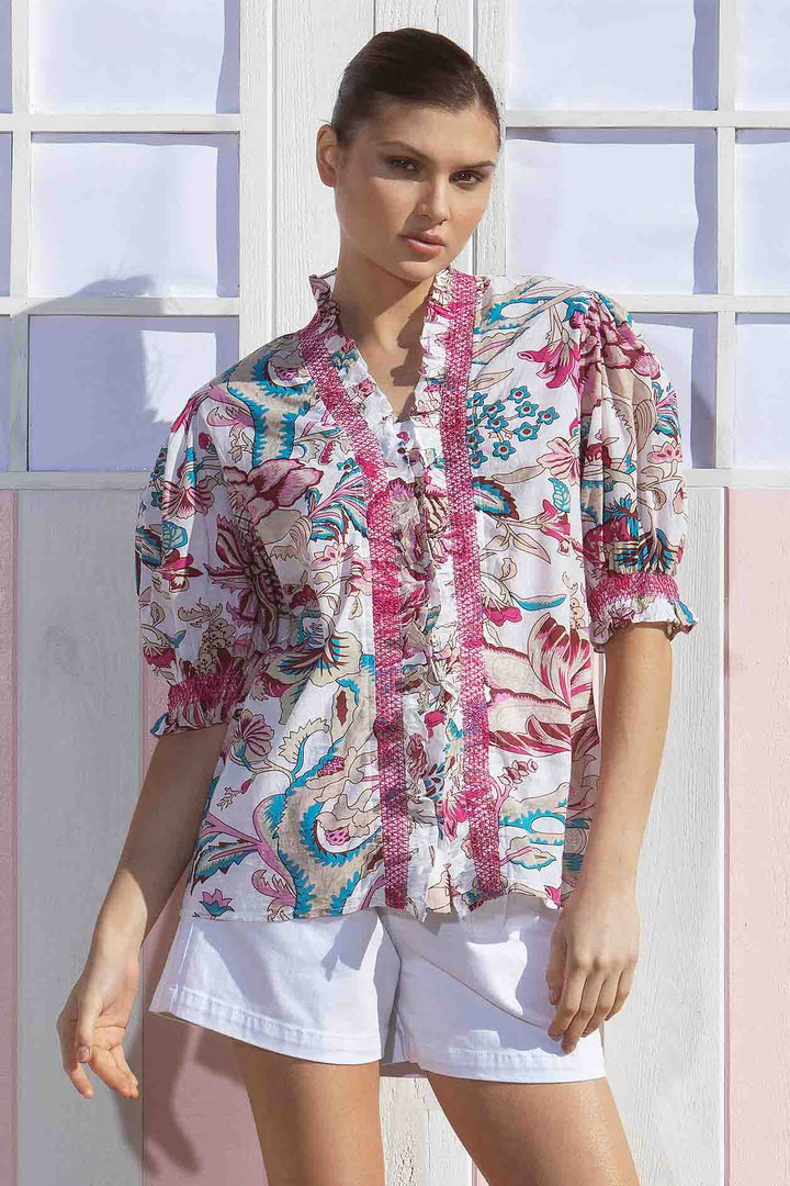 A model wearing a multicolored floral shirt with a flared collar and tapered sleeves, paired with white shorts, standing in front of a pink and white cabana.