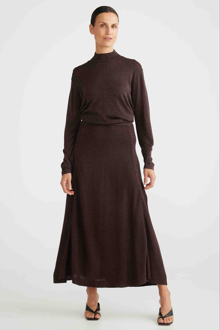 A woman wearing a dark cocoa colored knit skirt with a rib and seam detailing, paired with a matching top. The skirt reaches down to her ankles and has an elasticated waistband.