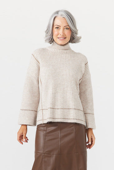 A woman wearing a beige turtleneck sweater with flared sleeves and a brown knee-length skirt with side splits. She is also wearing boots and the setting is a plain white background.