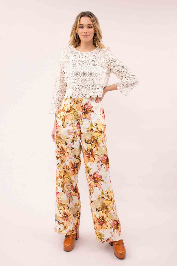 A model wearing a tropical floral print wide-legged pants with a fitted waist and front pleats. The pants are beige with a multicolor floral pattern.