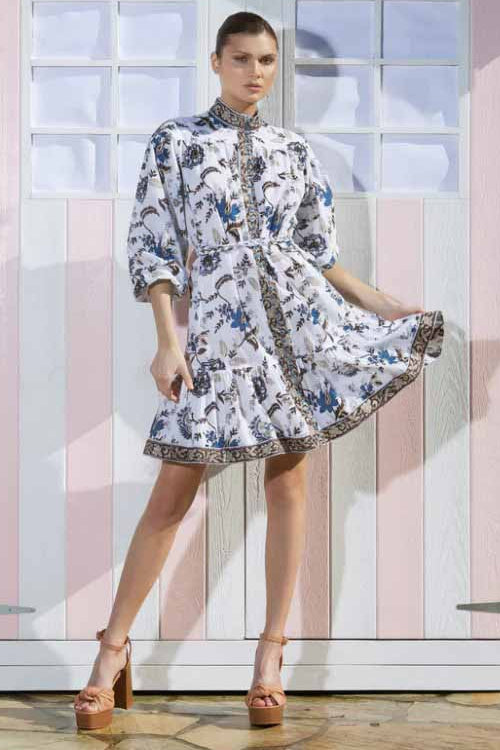 A model wearing a blue and white floral dress with long sleeves and a matching plaited belt, standing in front of a striped structure.