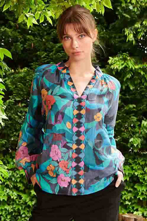 A person standing on a wooden deck wearing a floral pink and blue blouse with long sleeves, buttoned at the front, paired with black trousers. Accessory details include pink heels.