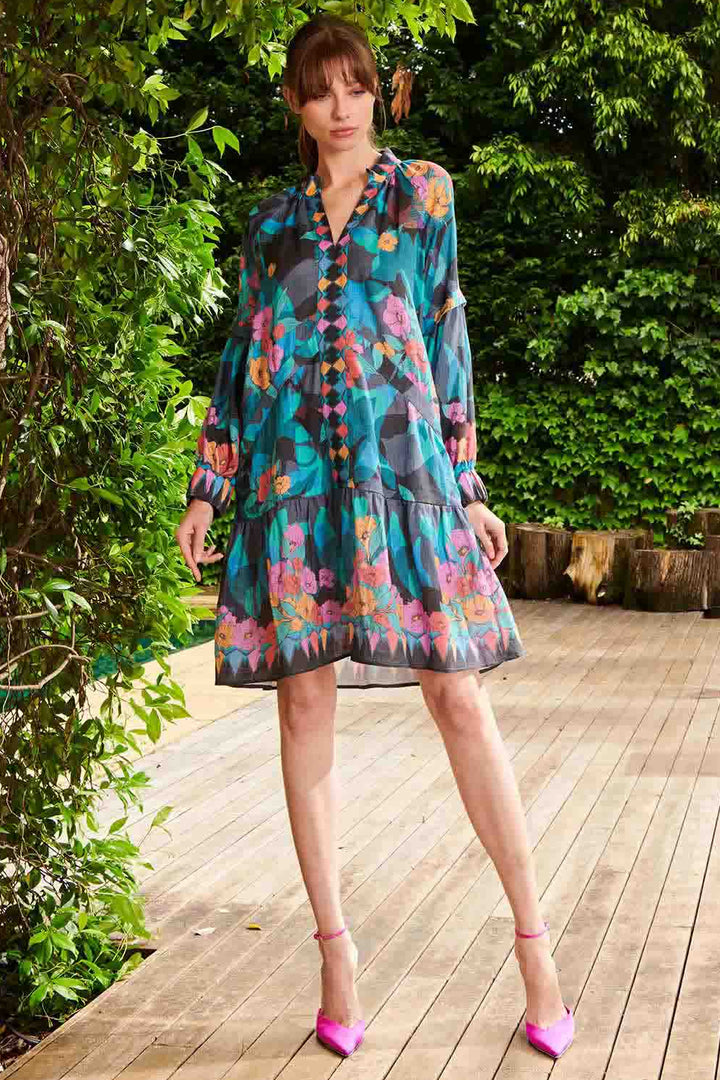 A woman standing on a wooden deck wearing a floral print, pink and blue, long sleeve mini dress with button details.