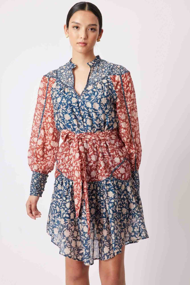 A model wearing a high neck, knee-length dress with a floral pattern in shades of blue and red. The dress features soft blouson sleeves and a removable waist tie.