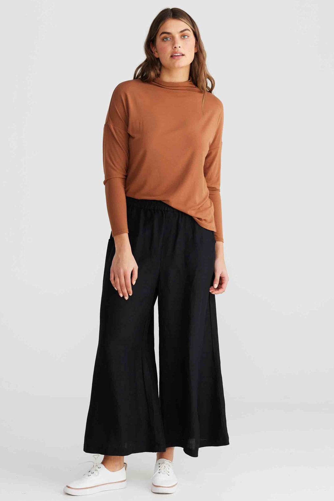 A woman modeling a rust-brown mock neck long-sleeve top and black wide-leg pants with white sneakers, photographed against a light gray studio background.