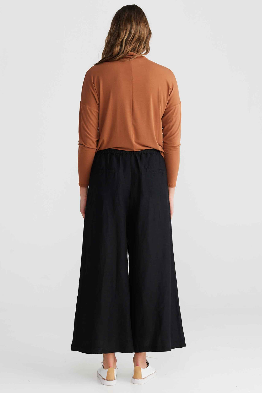 A woman viewed from the back wearing a rust-brown long-sleeve top and wide-leg black pants, standing against a light gray studio background.