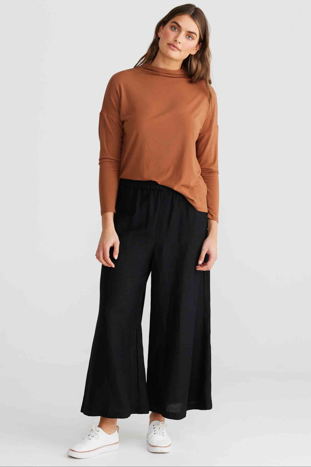 A woman modeling a rust-brown long-sleeve mock neck top and black wide-leg pants with white sneakers, photographed against a light gray studio background.
