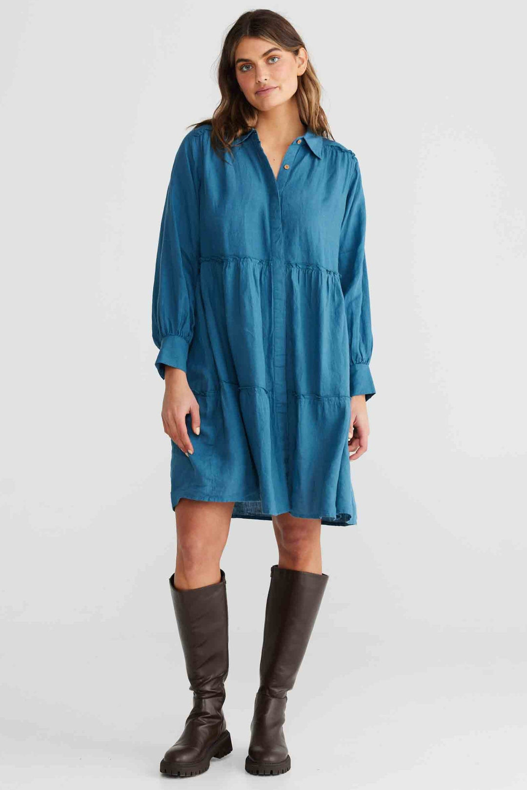 A woman standing in a studio setting, wearing a blue collared dress with long sleeves and a knee-length cut. She has accessorized with boots and jewelry.