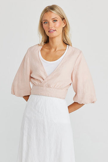 A woman wearing a peony linen wrap bolero with a V-neck front and waist length, paired with a white skirt. She is standing against a white background.