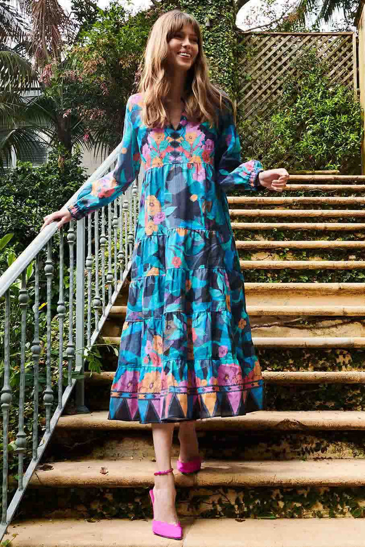 A woman standing on outdoor stairs wearing a floral Serena Elm dress in pink and blue tones with long sleeves and a tiered design.