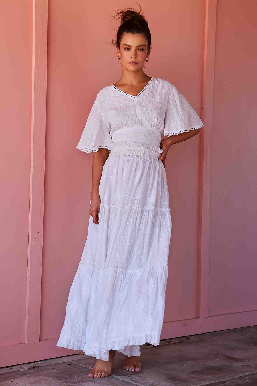 A woman wearing a white tiered maxi dress with short flutter sleeves and a smocked waist, standing barefoot against a pink wall.