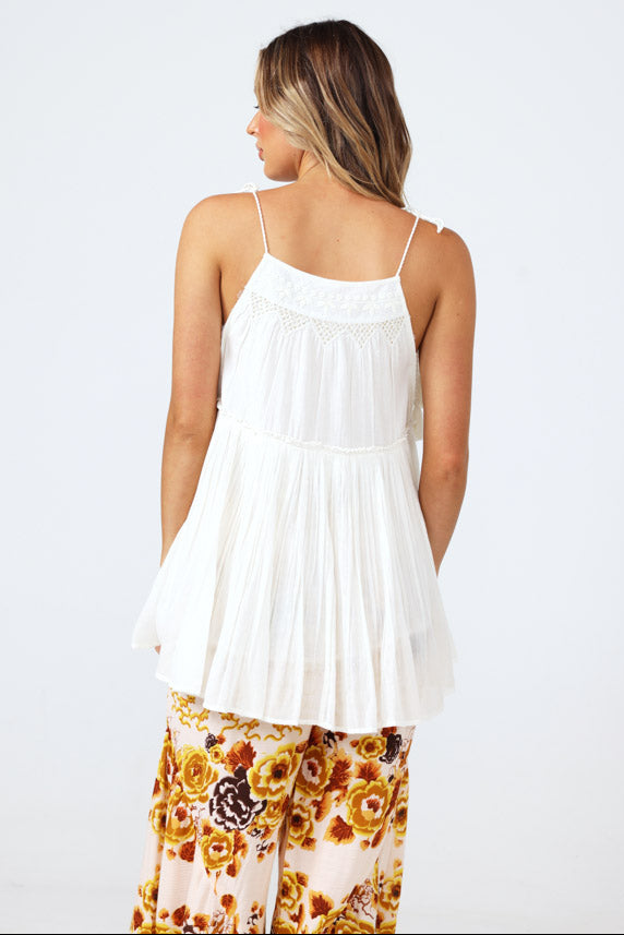 A woman shown from the back wearing a white sleeveless camisole-style top with thin straps and a flared gathered skirt, paired with floral pants, against a plain light gray studio background.
